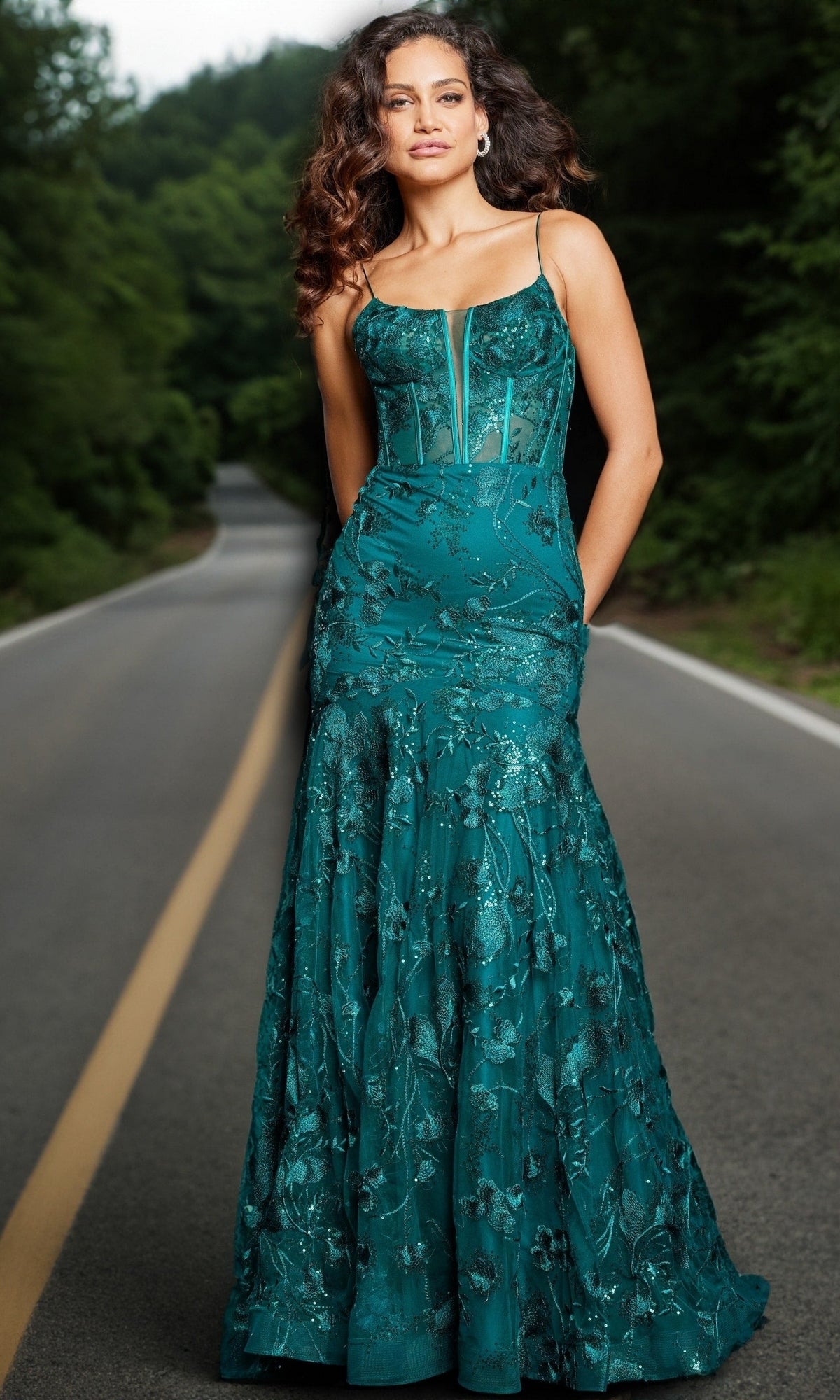 A woman with wavy hair stands on a road in the Jovani 38004 long corset mermaid prom dress, featuring thin straps and a floral pattern, surrounded by greenery.